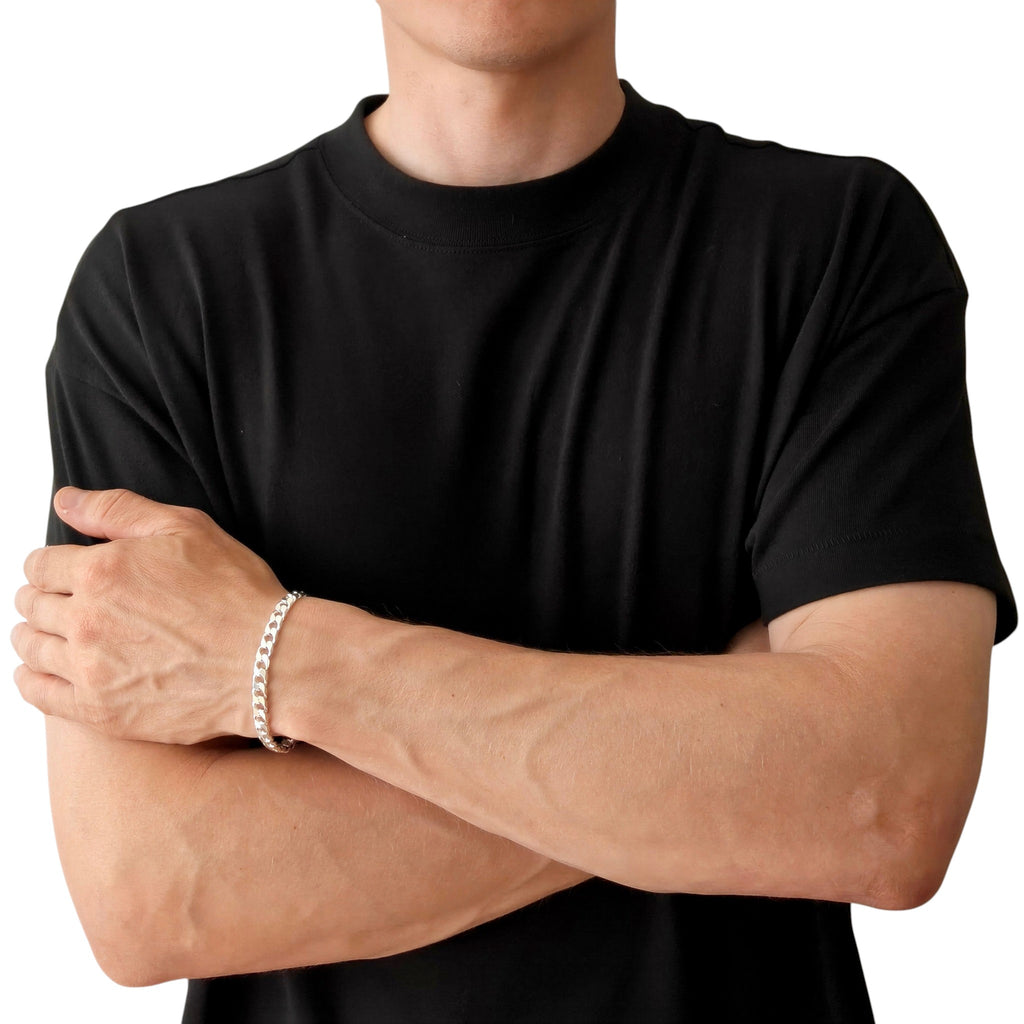 Neviera curb link chain bracelet — Italian-made 925 sterling silver, 6,4mm width. Lifestyle photograph on model wearing the bracelet with a black crew-neck t-shirt on a neutral background, alternate pose. Neviera specializes in high-quality Italian-made 925 silver chain necklaces and bracelets.