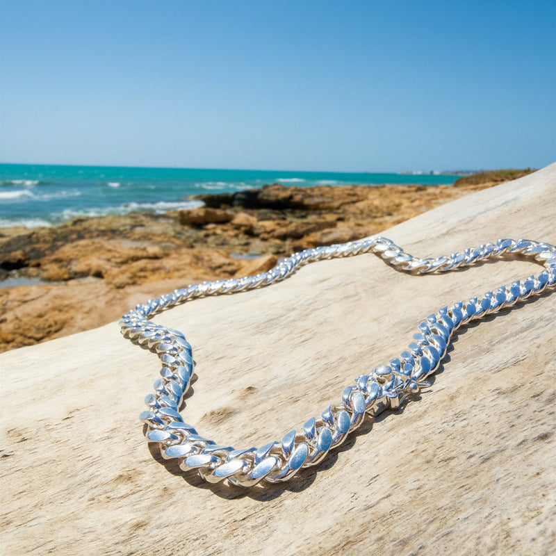 A chunky Neviera silver Cuban link chain necklace draped over a light-colored wooden log with a rocky beach and blue ocean in the background.